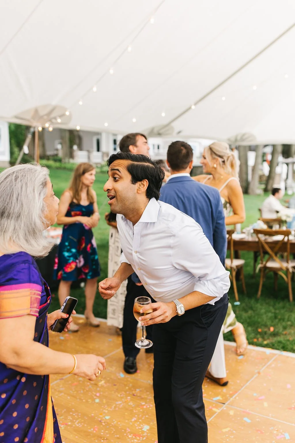 People socializing at an outdoor celebration under a large tent, with a man in a white shirt and black pants holding a glass of rosé wine engaged in conversation with an older woman wearing a colorful dress.