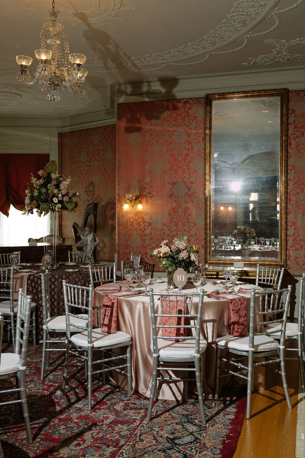 Elegant dining room with round table set with pink tablecloth, floral centerpiece, glassware, and silver chairs. Vintage decor includes a chandelier, ornate mirror, and classical statue with harp in the corner. Patriotic red and gold wallpaper and a 