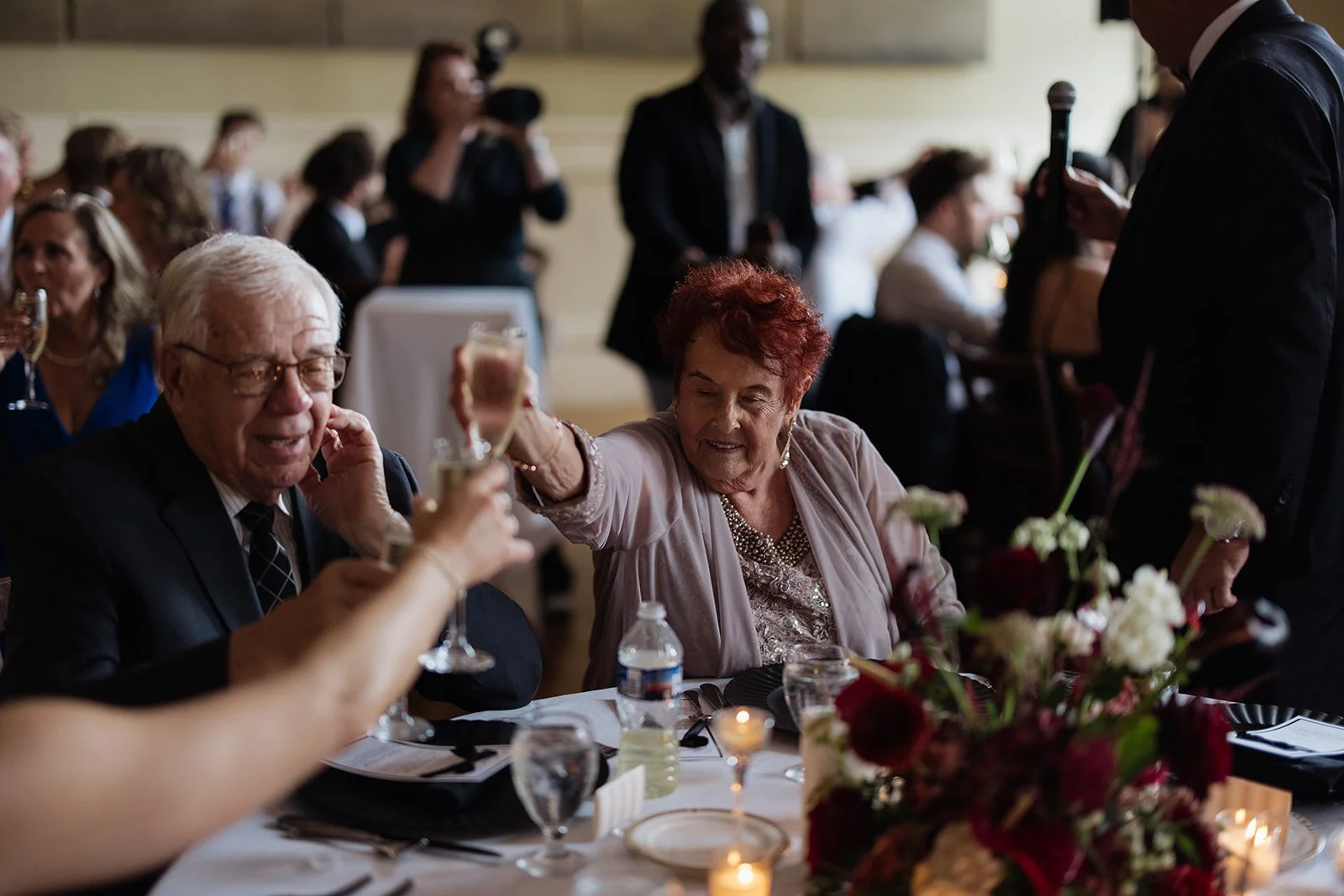 Elderly man and woman celebrating with glasses of champagne at a festive table, surrounded by other guests at a social event.