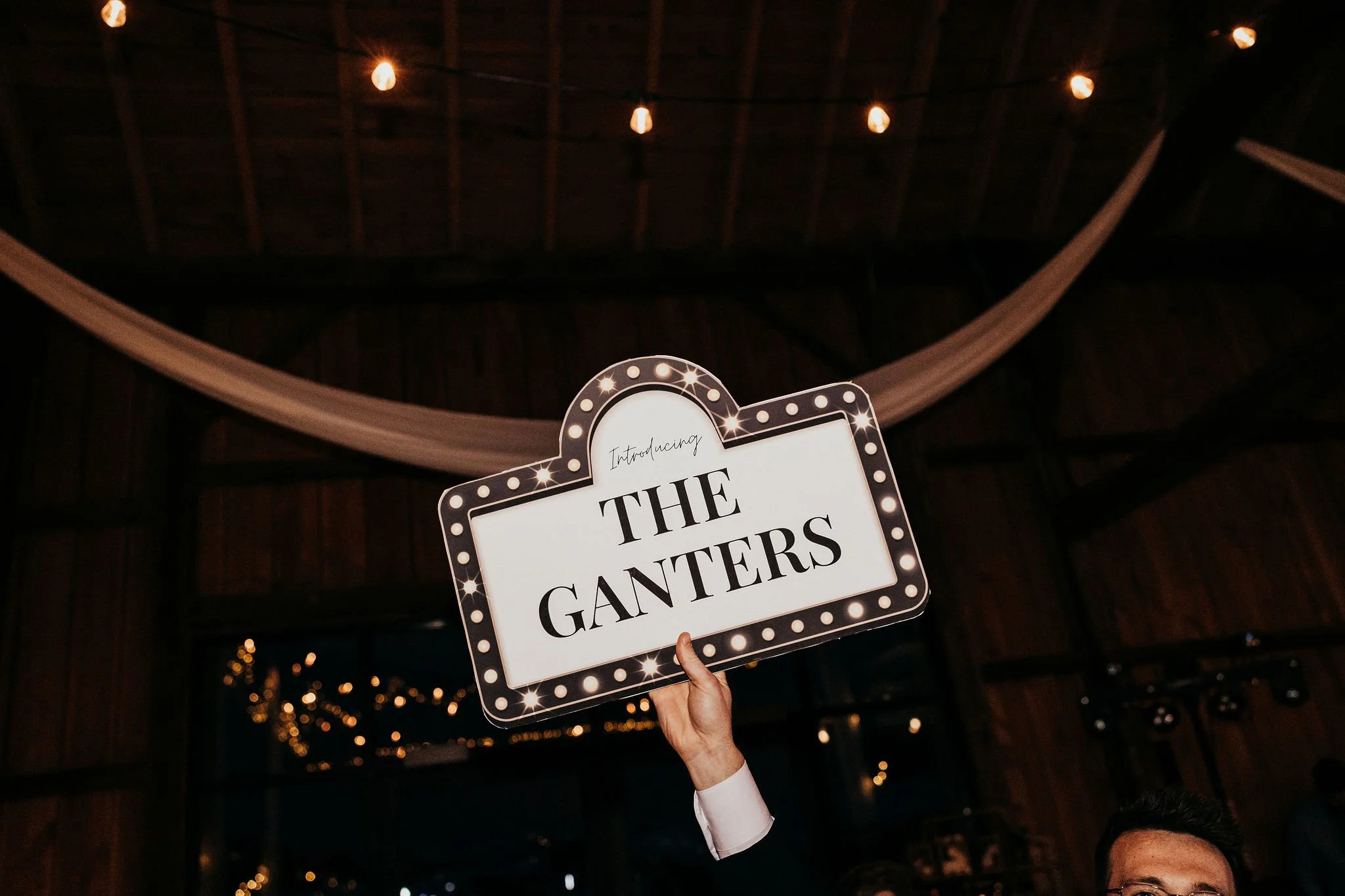 A person holding a sign that reads "Introducing THE CANTERS" in a festive setting with string lights and a wooden ceiling.