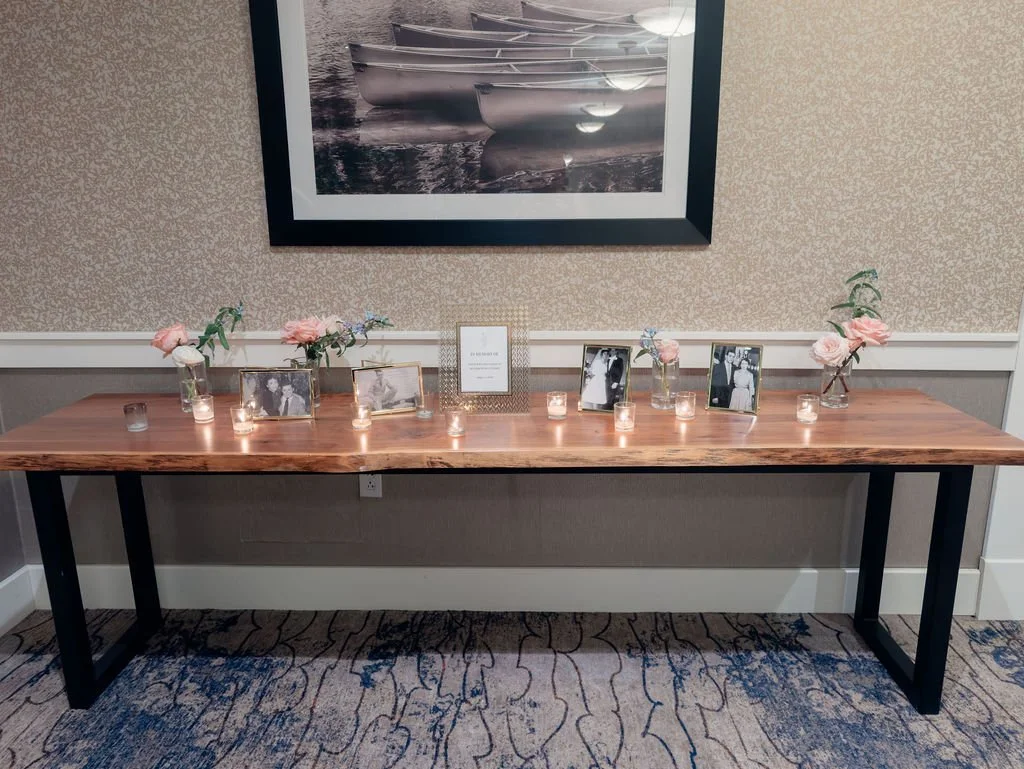 A wooden table decorated with framed photos, vases with pink and white flowers, and lit candles, set against a wall with a large framed black and white picture.