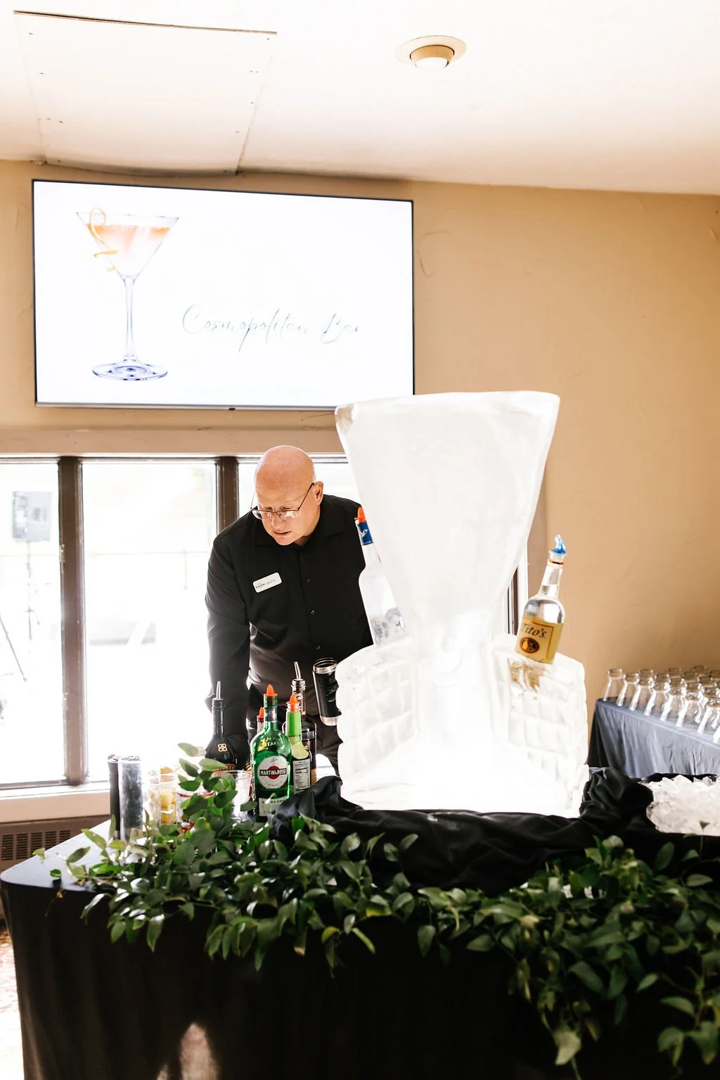 Bartender preparing drinks at a bar with a large ice sculpture that looks like a cocktail glass. A screen in the background displays a martini glass and the words 'Cosmopolitan Bar.'