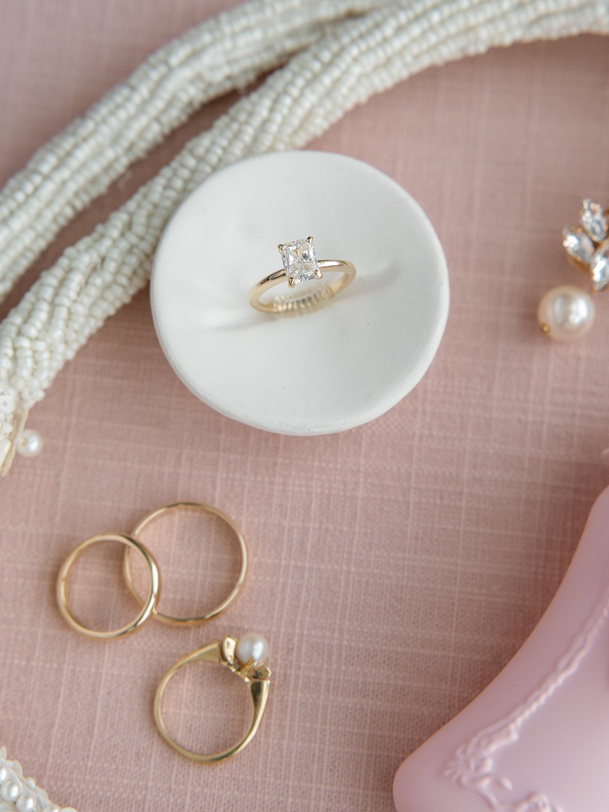 A close-up of a gold ring with a large square-cut diamond on a white stand, surrounded by pearl necklaces, gold rings, a pearl earring, and decorative jewelry pieces on a pink surface.
