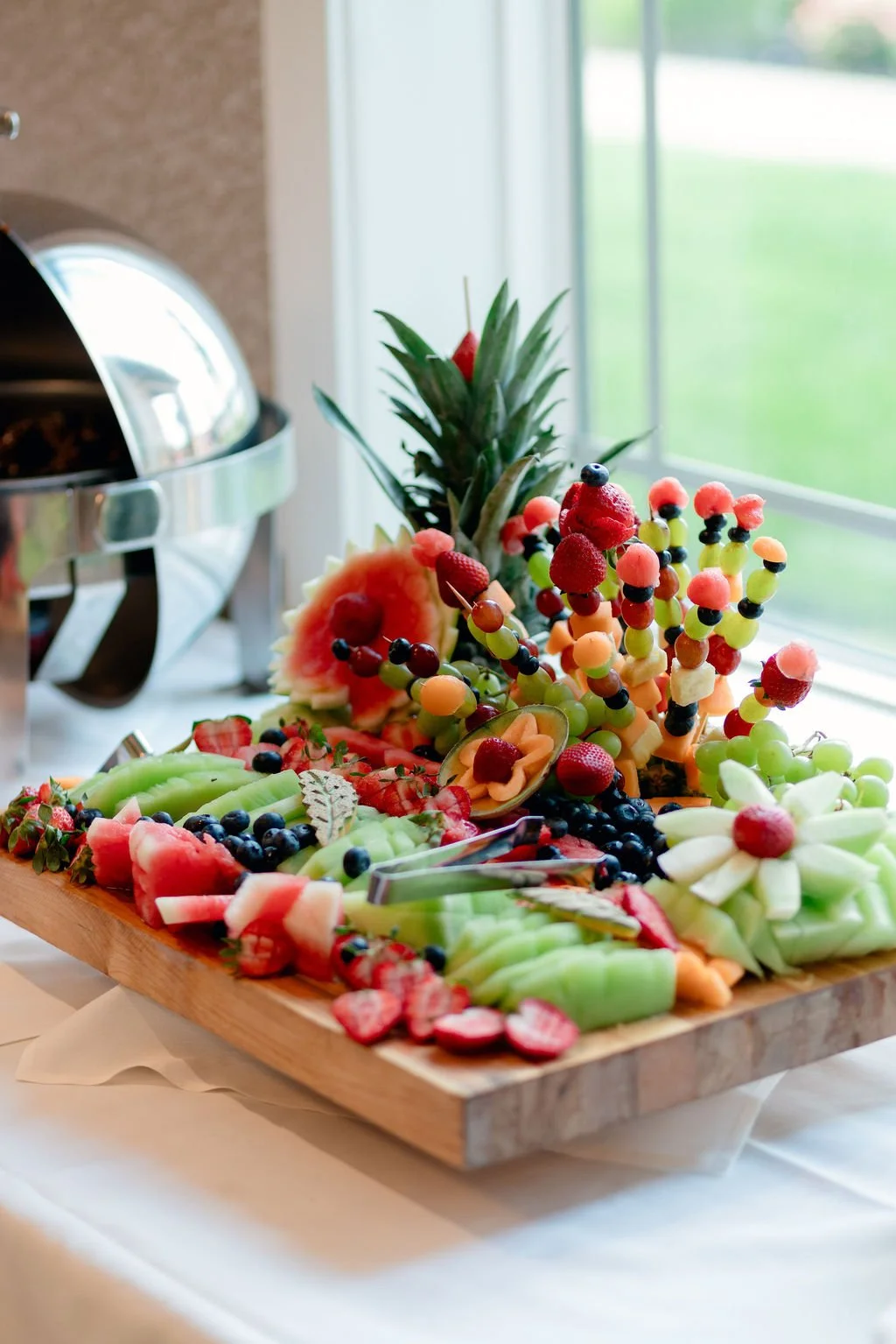 A wooden platter with assorted fresh fruit arranged around a pineapple, including strawberries, blueberries, grapes, cantaloupe, watermelon, honeydew, and carved melon flowers, placed on a white table near a window.