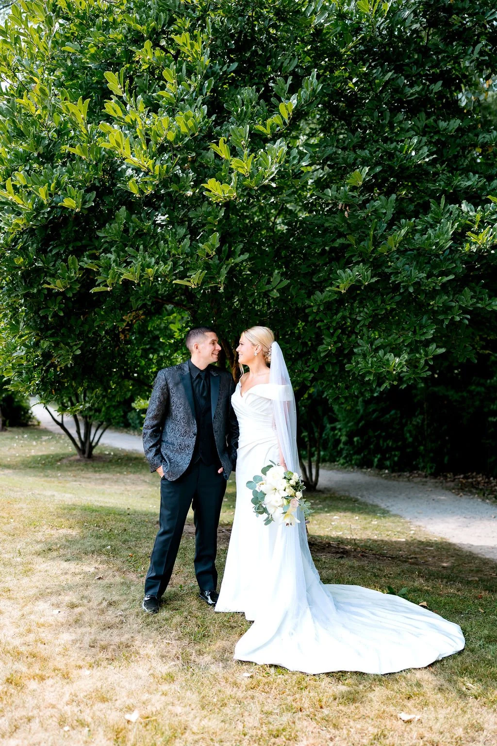 A bride and groom stand together outdoors beneath a large leafy tree, looking at each other and smiling. The bride wears a white wedding gown with a long train and veil, holding a bouquet of white flowers and greenery. The groom wears a black suit wi
