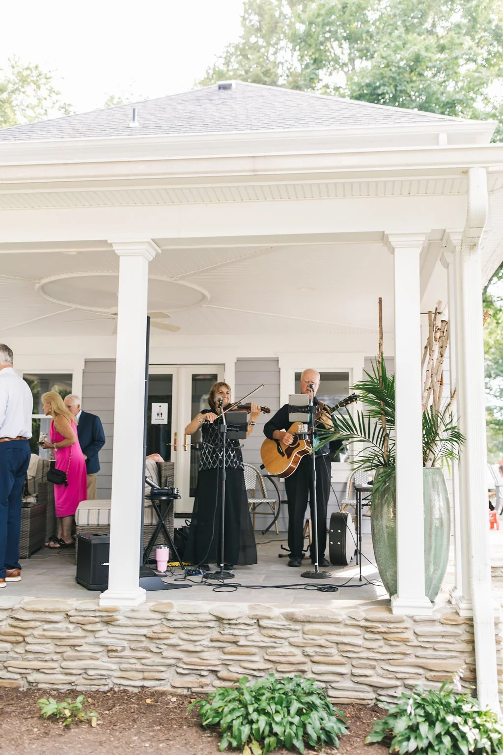 Musicians performing on an outdoor patio with a singer playing a violin and a guitarist, surrounded by guests and plants, during daytime.