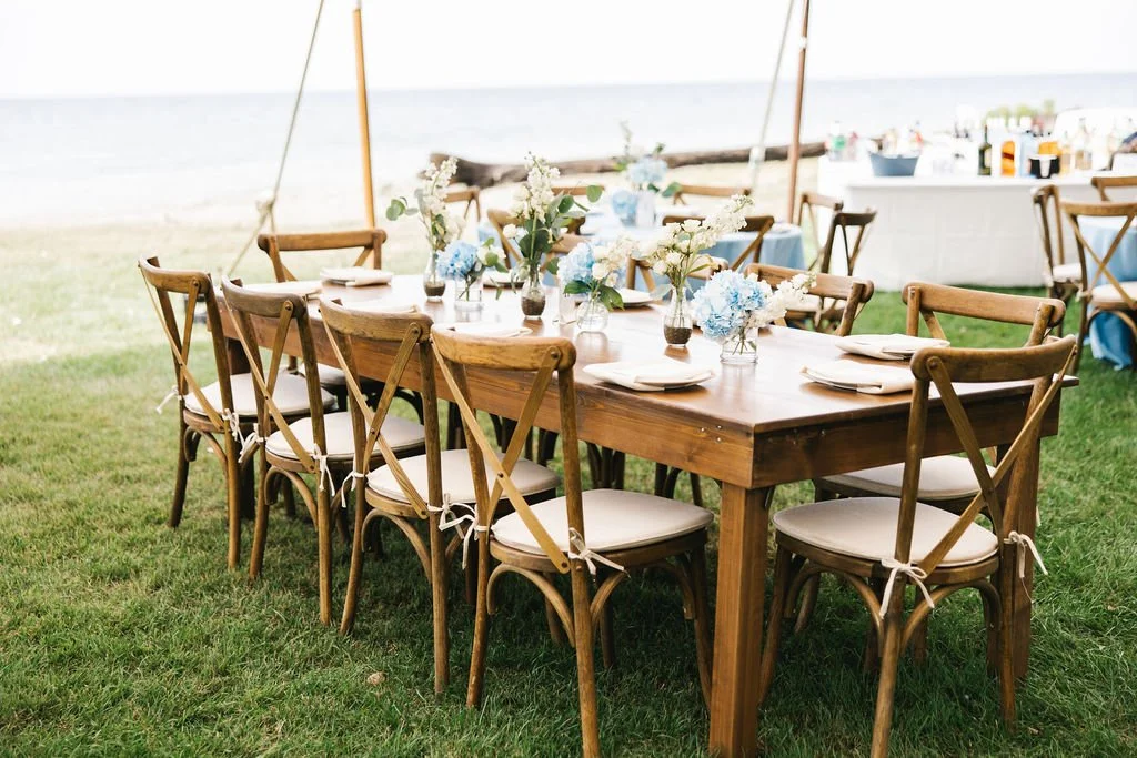 Outdoor dining table set for a celebration with floral centerpieces, on a grassy area near the beach.