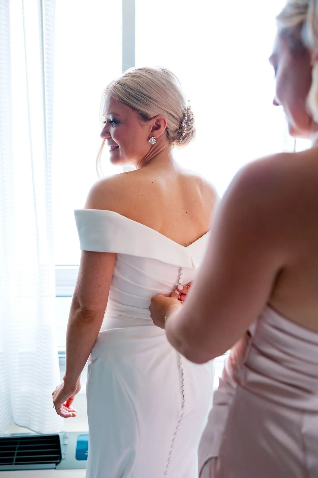 A bride in a white wedding gown being assisted with the back buttons by another woman, standing in front of a window with white curtains.