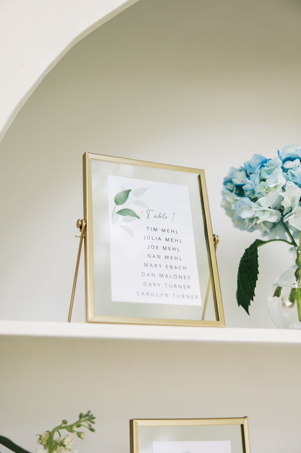 A framed seating chart with a list of names in front of a bouquet of blue and white hydrangea flowers in a glass vase on a white wall shelf.