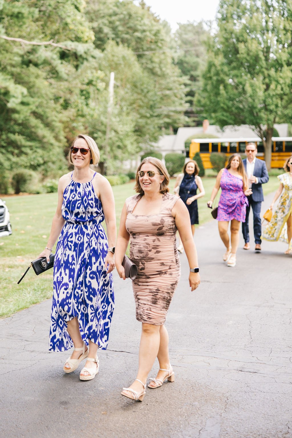A group of women dressed in summer attire walking outdoors on a paved path, smiling and enjoying themselves, with trees and a yellow school bus in the background.