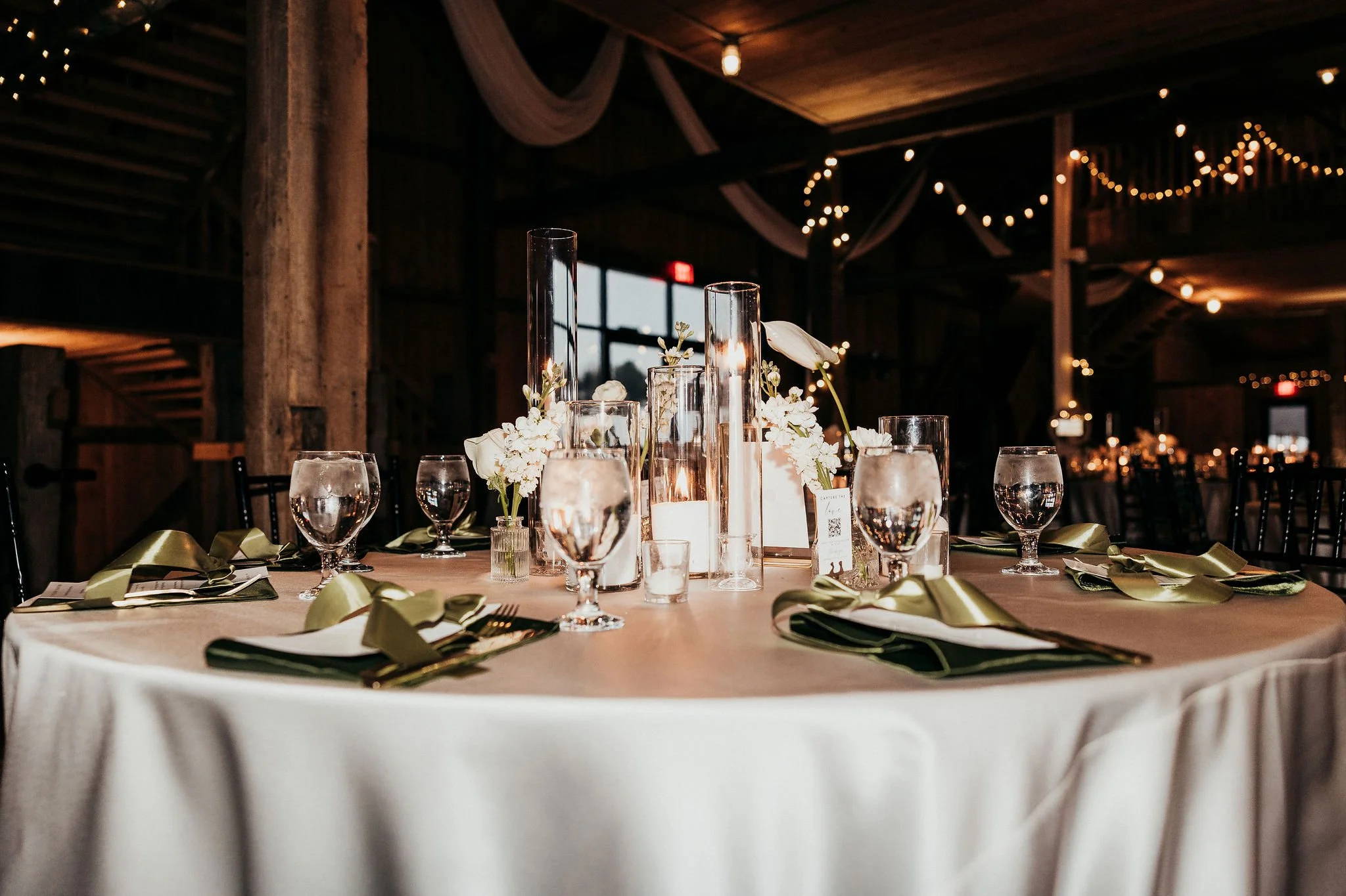 A round banquet table with a white tablecloth, set with water glasses, gold-colored napkins, and floral centerpieces in tall glass vases, in a dimly lit rustic venue decorated with string lights and draped fabric.