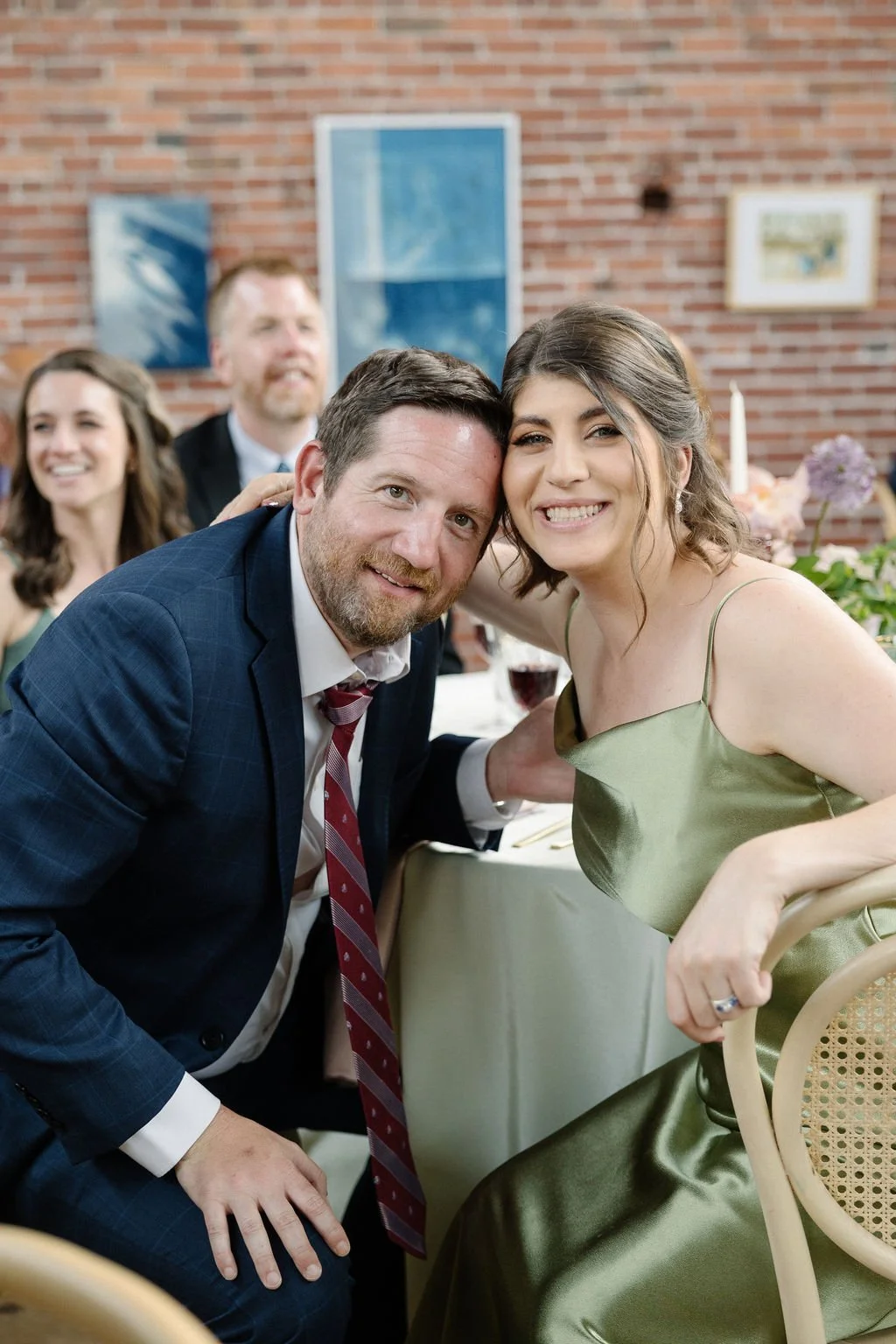 A group of people at a celebration or social event, with a focus on a smiling woman in a green dress and a man in a dark suit kneeling beside her, both leaning in close for a photo.