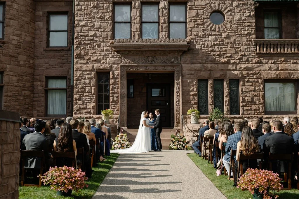 A wedding ceremony with the bride and groom holding hands in front of an audience, outside a large stone building decorated with flowers and greenery.