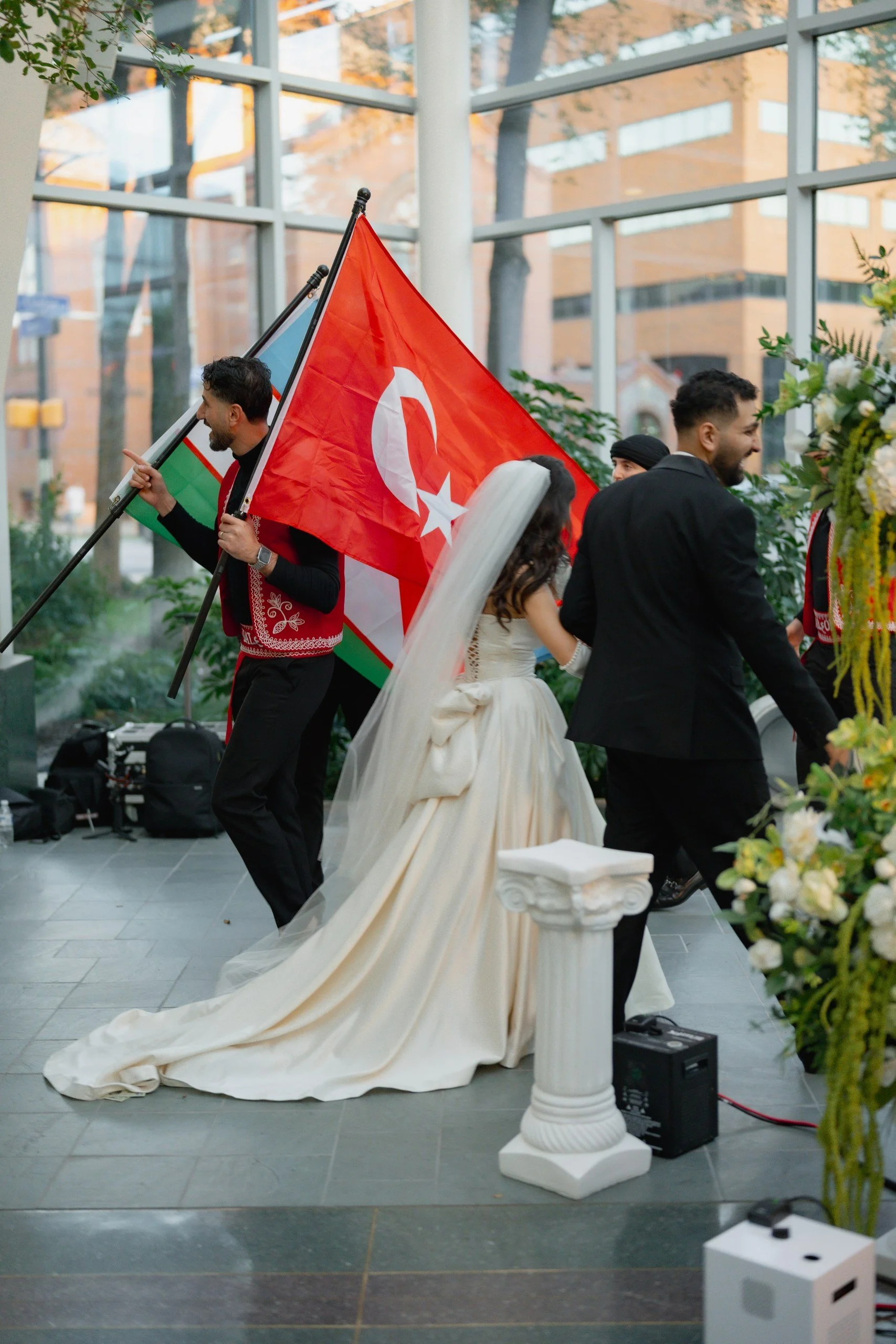 People participating in a wedding celebration inside a building with large glass windows, holding Turkish flags and wearing formal and traditional attire.