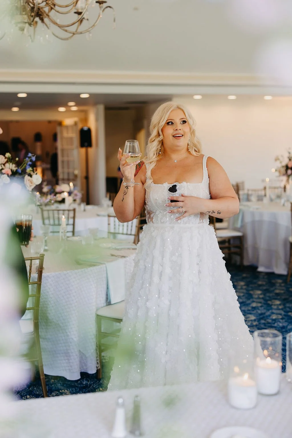 A woman in a white wedding dress holding a glass of white wine and a small glass with a dark drink, standing in a decorated reception hall with tables, floral centerpieces, and candlelit decor.