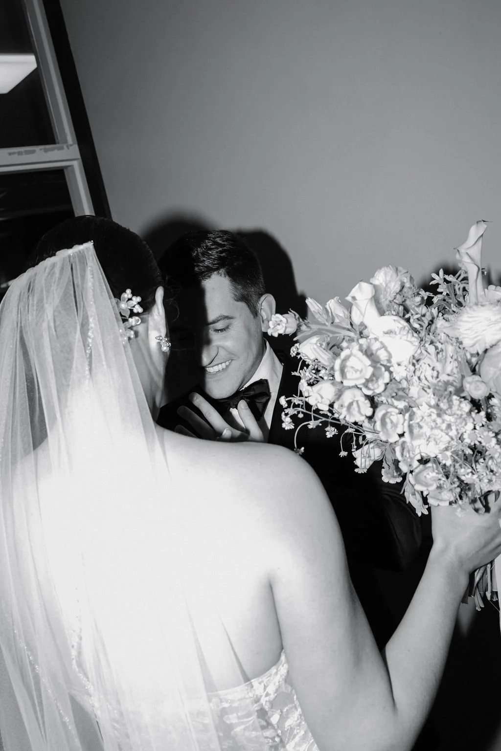A black and white photo of a bride and groom sharing a joyful moment, with the bride holding a large bouquet of flowers and the groom smiling with his eyes closed.