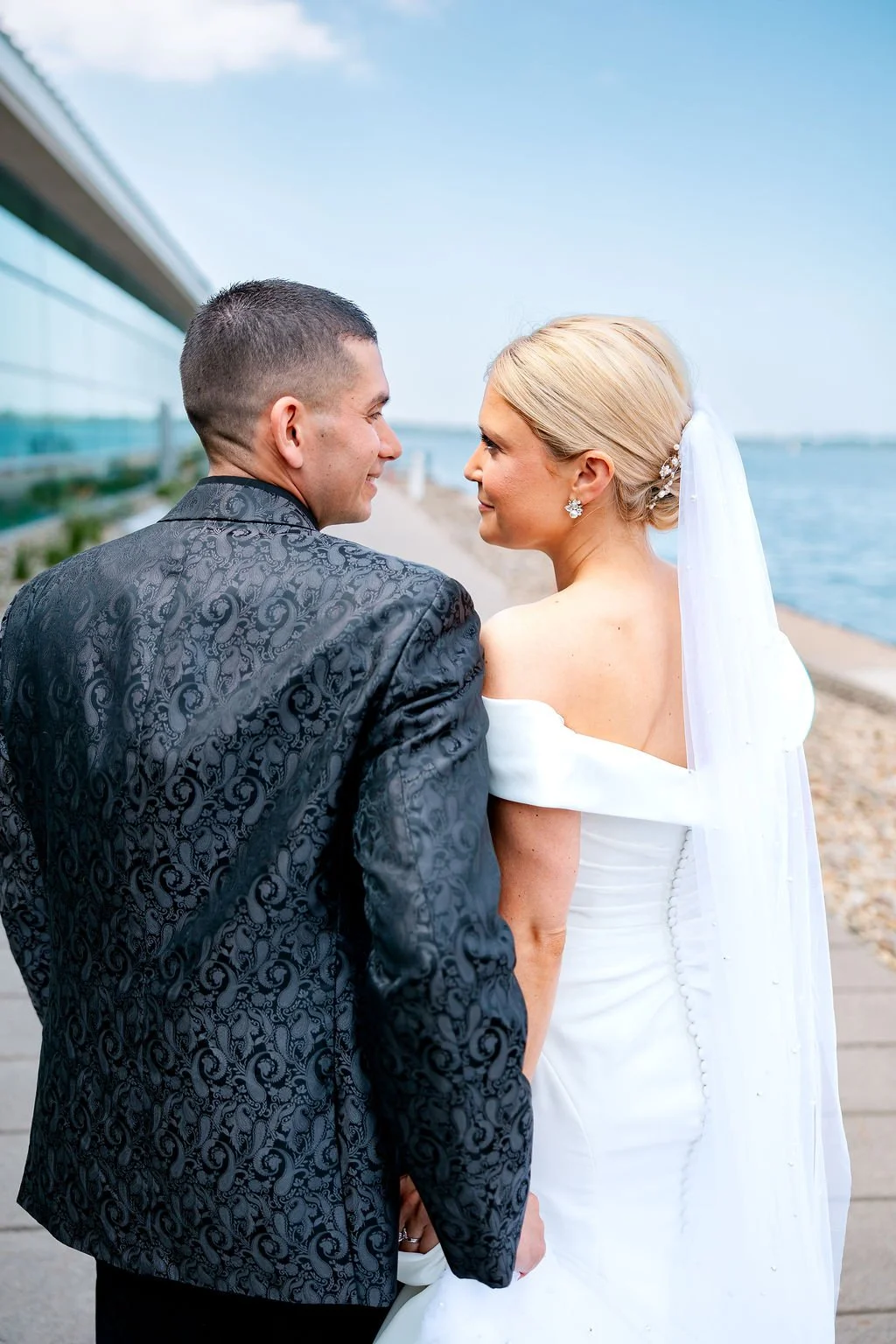 A bride and groom stand close together on a waterfront promenade, smiling at each other against a backdrop of water and sky, with modern buildings reflecting in the glass windows behind them.