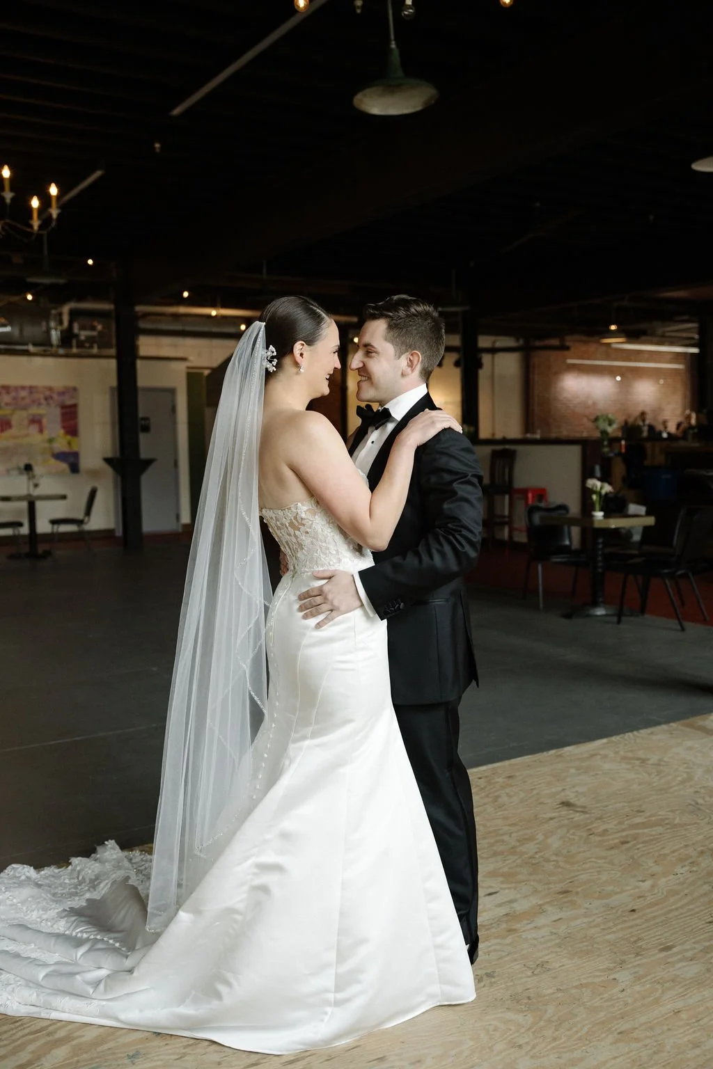 A bride and groom dance closely at their wedding reception in an industrial-style venue, with dim lighting and rustic decor.