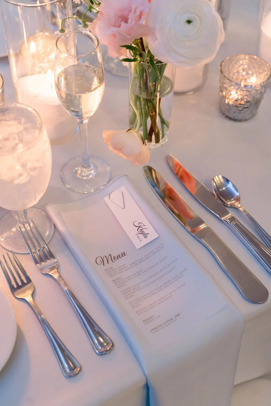 A table setting with a menu, silverware, glasses of water and champagne, a floral centerpiece with pink and white flowers, and candles in silver holders, all in an elegant setting.
