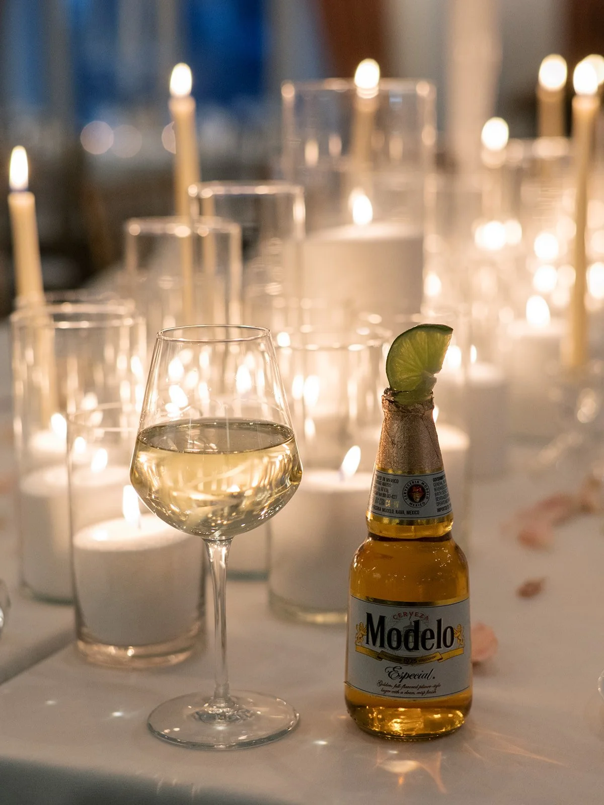 A glass of white wine, a Modelo beer with lime wedge, and lit candles in the background on a table, likely at a celebration or romantic setting.