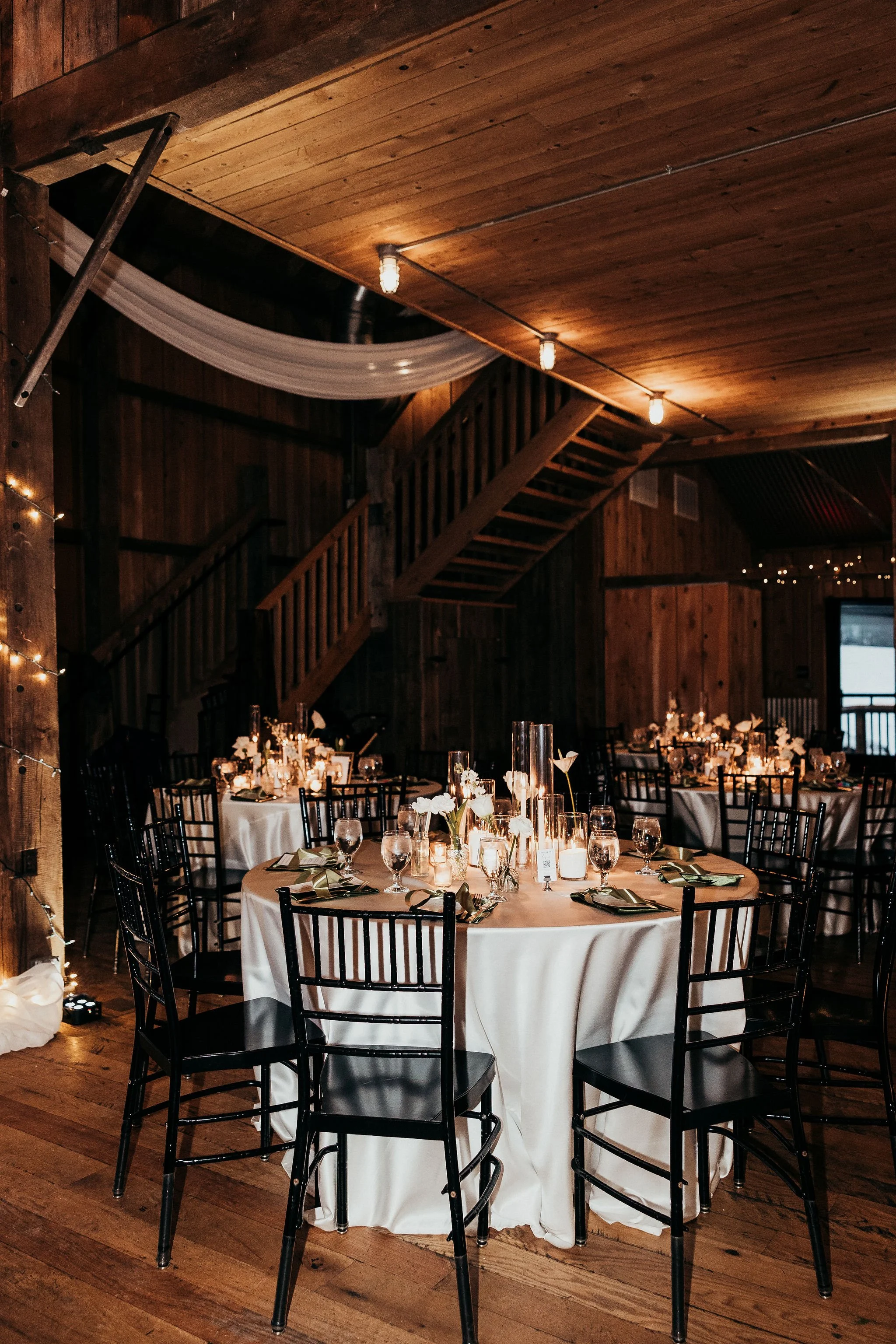 Indoor event space with round tables draped in white tablecloths, decorated with candle centerpieces and floral arrangements, surrounded by black chairs. The room features wooden walls, a wooden ceiling with string lights, and a staircase in the back
