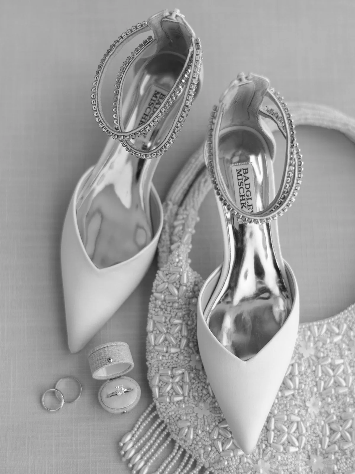 Elegant bridal shoes with intricate accents, jewelry, and rings displayed on a textured surface.