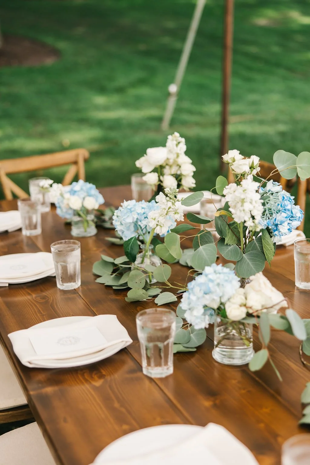 A decorated outdoor dining table with white and light blue floral centerpieces, clear glass drinking glasses filled with water, and white napkins on white plates, set on a wooden table. Green grass is visible in the background.