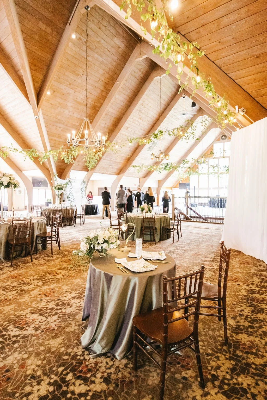 Wedding reception in a spacious indoor venue with wooden vaulted ceilings, decorated with hanging greenery and string lights. Round tables with floral centerpieces and chairs are arranged around the room, and a few people are seen standing and social