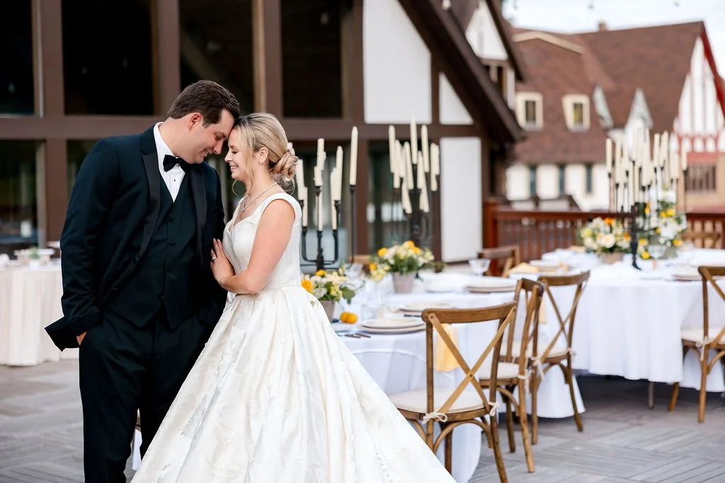 A bride and groom in wedding attire sharing a tender moment on an outdoor terrace with decorated tables in the background.