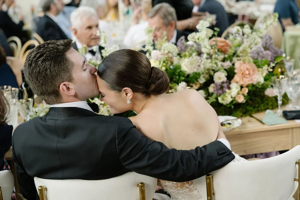 A newlywed couple at their wedding reception, with the groom kissing the bride on the forehead as she leans into him, surrounded by floral arrangements and seated guests.