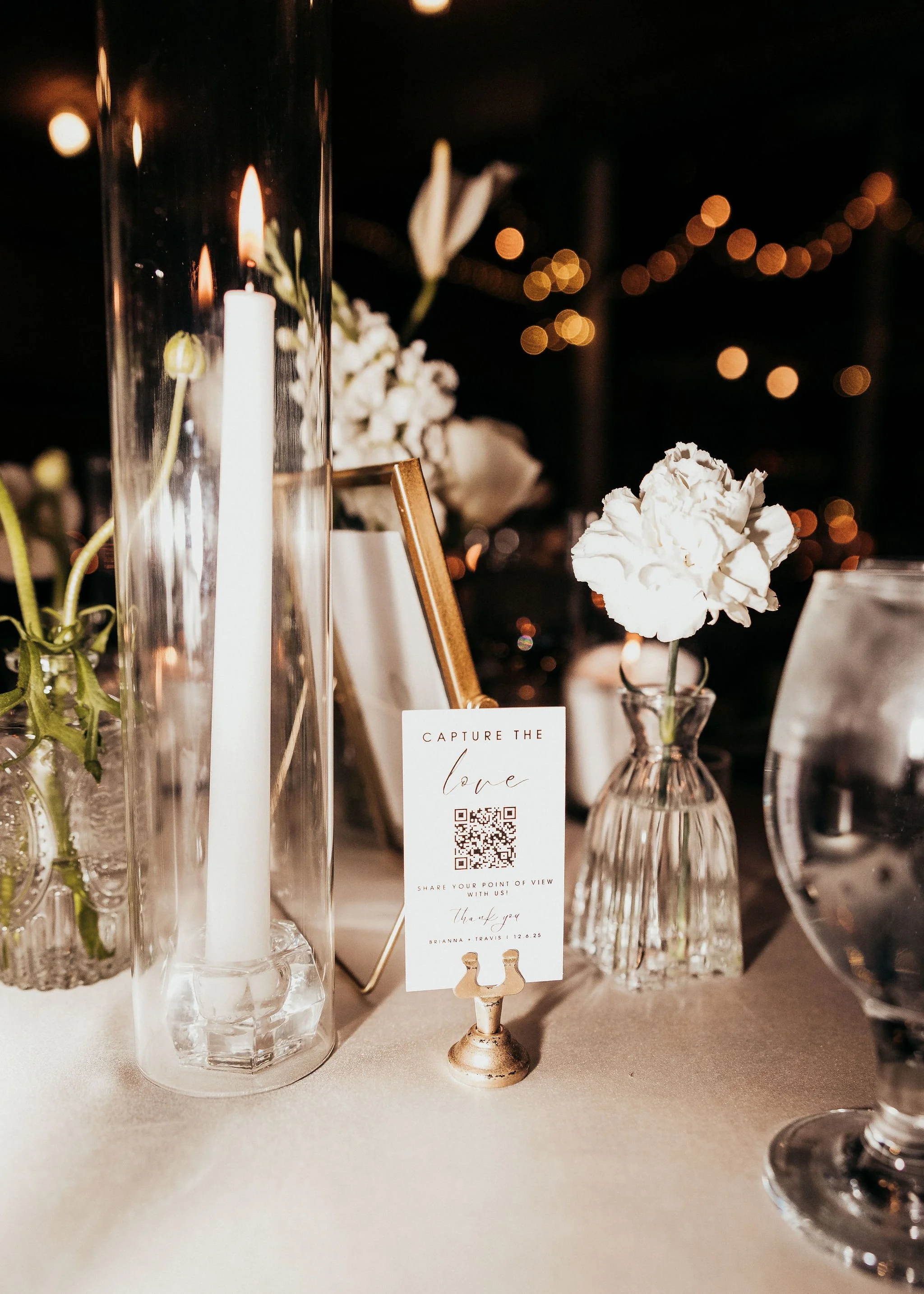 Close-up view of a wedding table centerpiece with white flowers in small glass vases, a tall glass candle holder with a white candle, and a table card with a QR code and a message to share the point of view. Warm bokeh lights in the background.