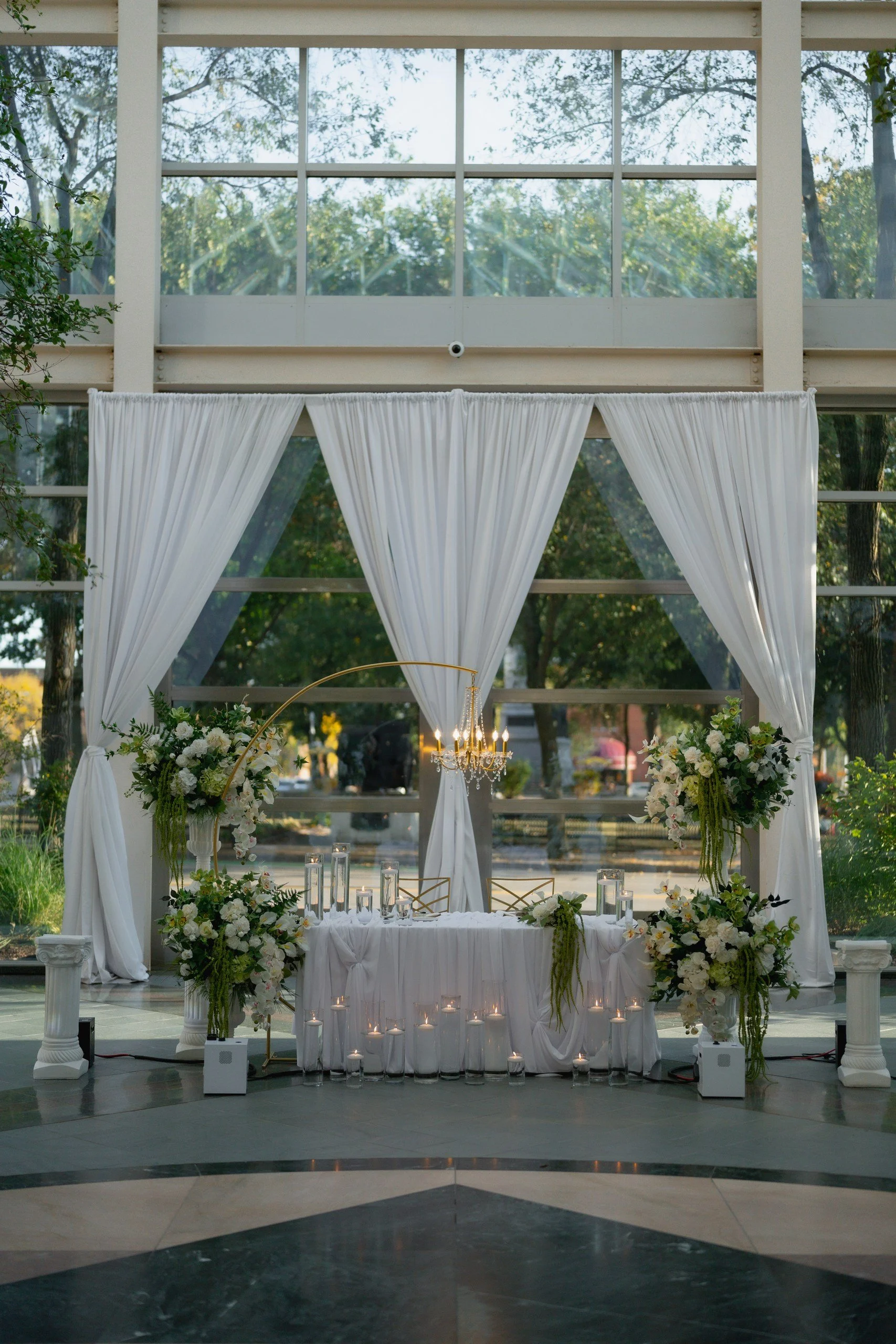 Elegant indoor wedding ceremony setup with white drapery, floral arrangements, and a chandelier, positioned in front of large windows with a view of trees.