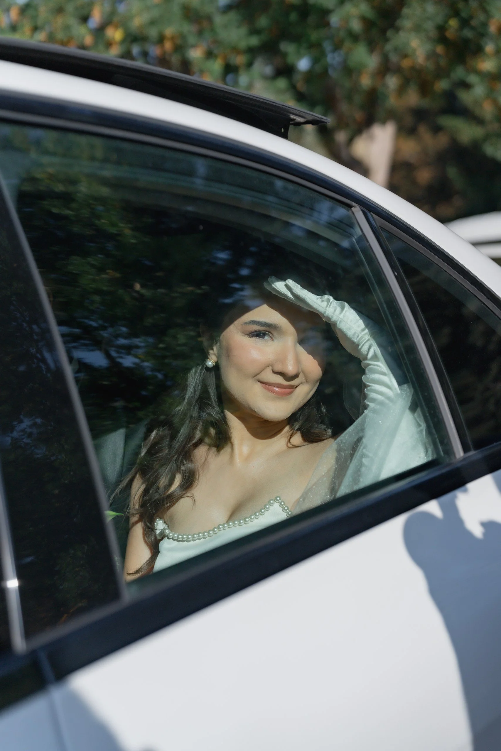 A woman with dark hair and pearl earrings, dressed in a white dress with pearl accents, sitting inside a car with reflections of trees on the window, smiling and saluting with her right hand.