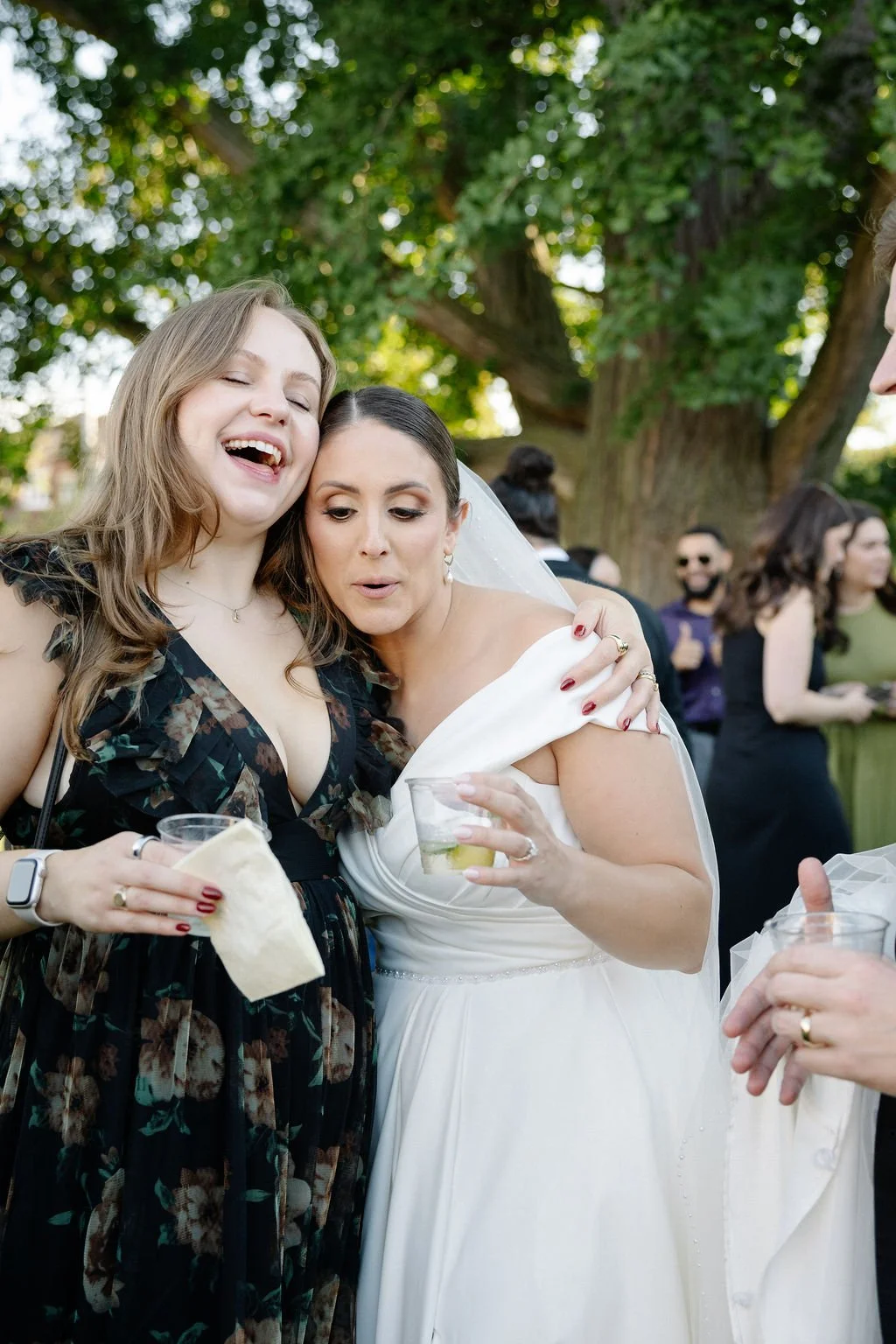 Two women, one in a wedding dress and the other in a dark floral dress, hugging and holding drinks at an outdoor gathering with trees and people in the background.