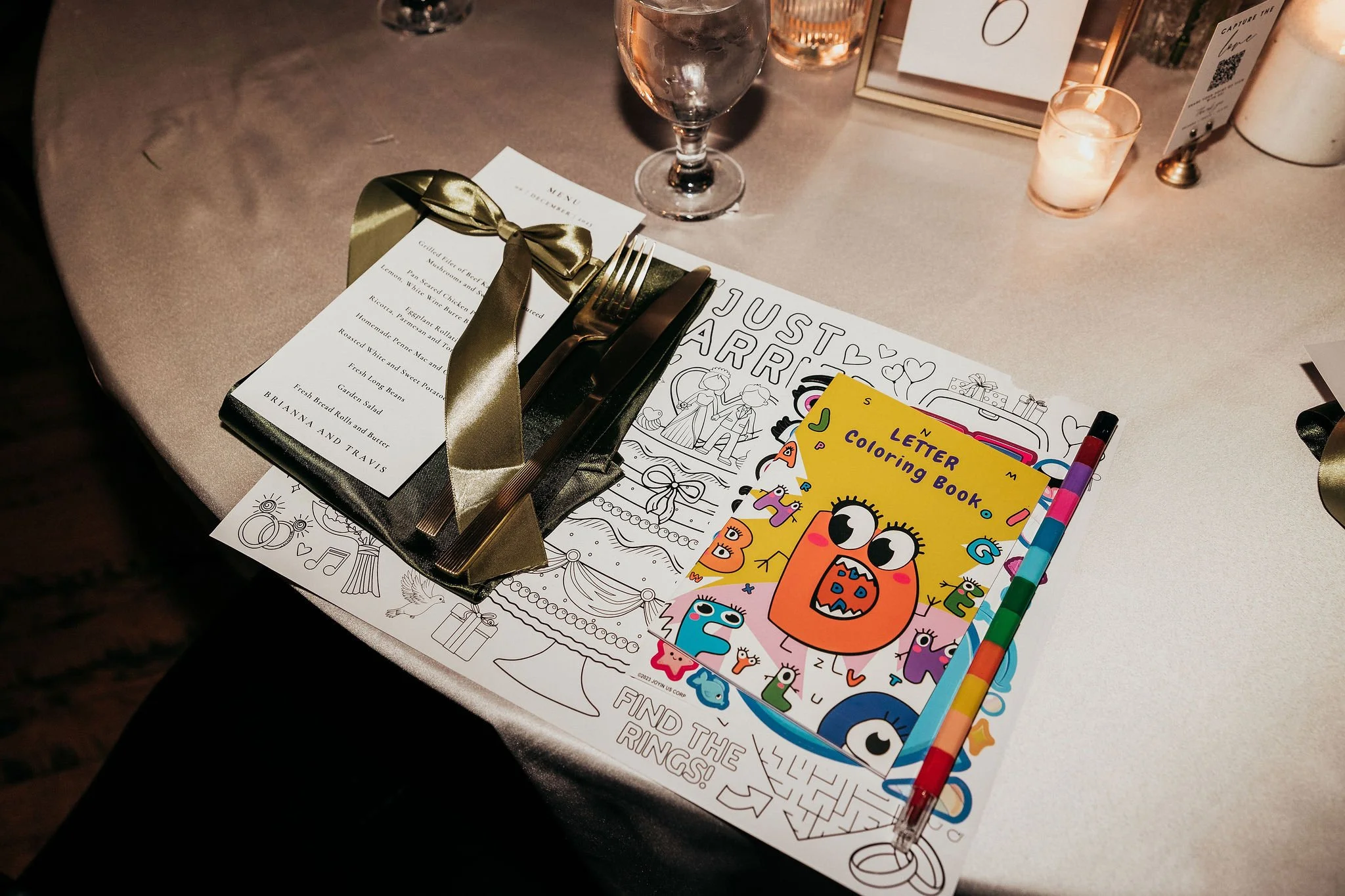 A table with several items, including a gold ribbon, a black-bordered menu, a fork and knife, a water glass, a candle, and children's activity and coloring books.