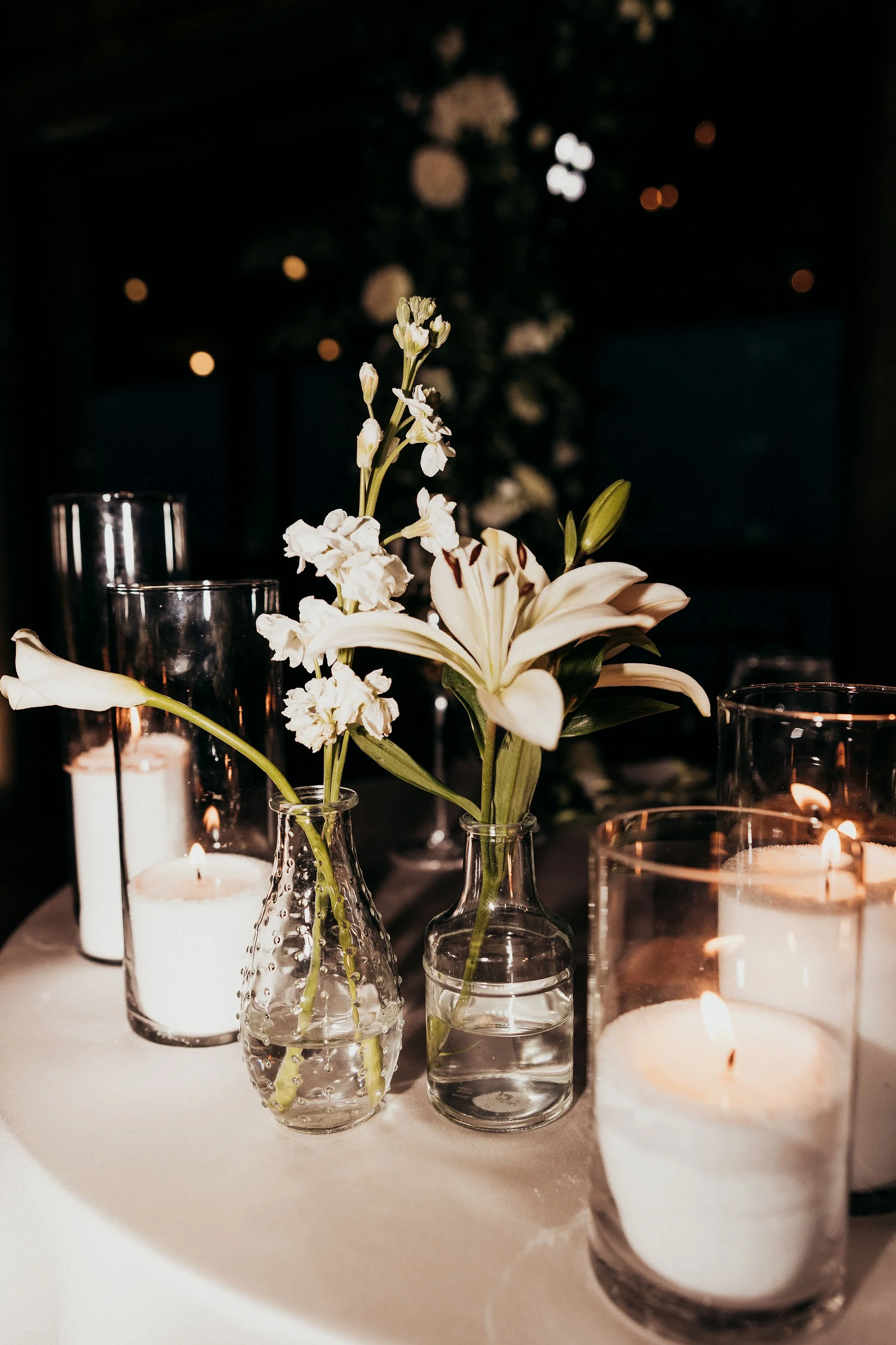 Vases with white flowers and lit candles on a table at night, blurred background with warm lights.