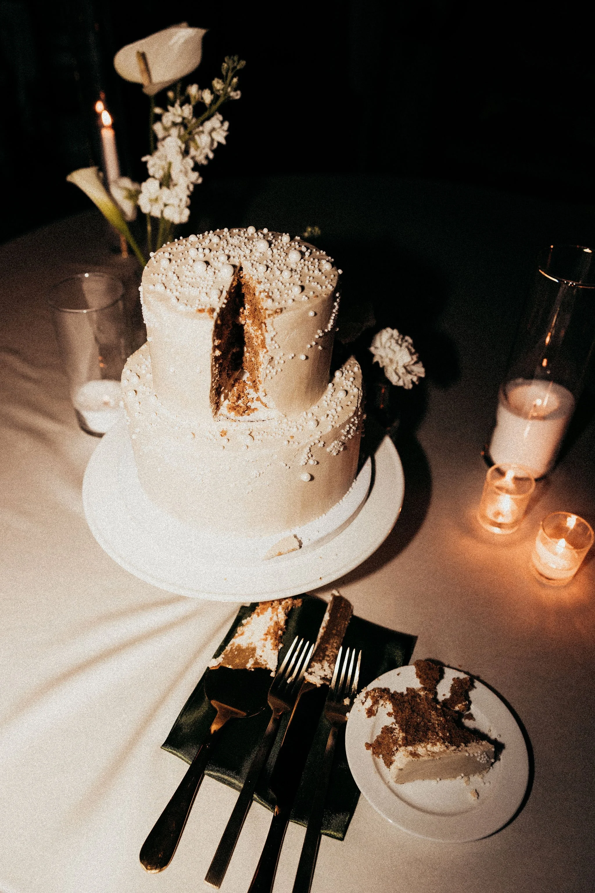 A partially sliced cake with white icing and pearl decorations on top, set on a white cake stand. There are flowers and candle holders with white candles surrounding the cake. A piece of cake on a plate with a fork is in the foreground.