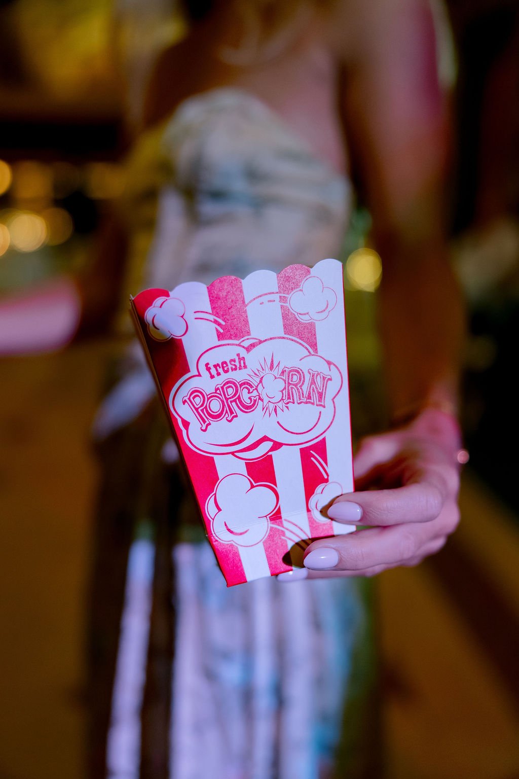 A person holding a small red and white striped box of fresh popcorn. The background is blurry with warm lighting.