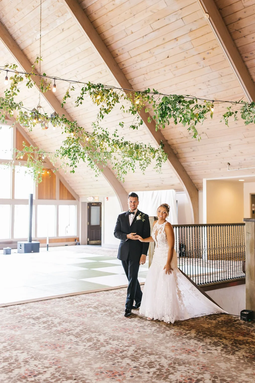A bride and groom standing together indoors at a wedding reception, smiling and holding hands, with greenery and string lights hanging from the wooden ceiling.