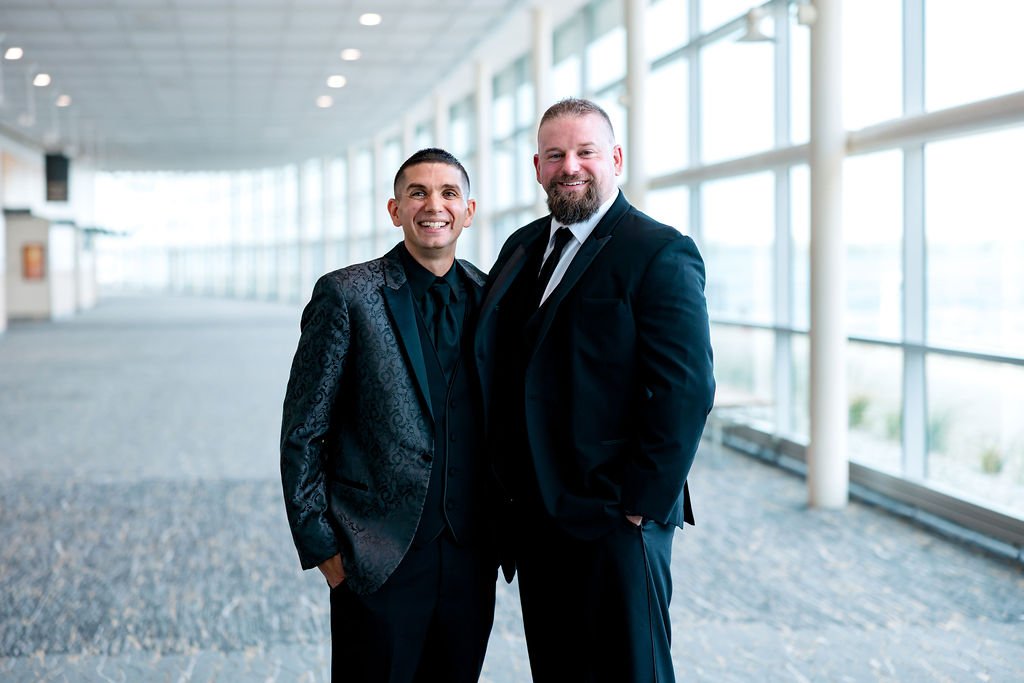 Two men in suits smiling at the camera, standing in a bright, modern office or conference center with large windows and natural light.