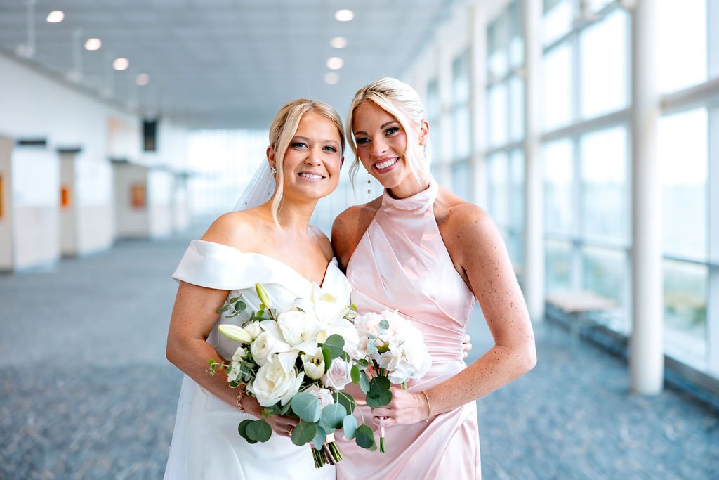 Two women in wedding attire smiling and holding bouquets inside a modern building with large windows.