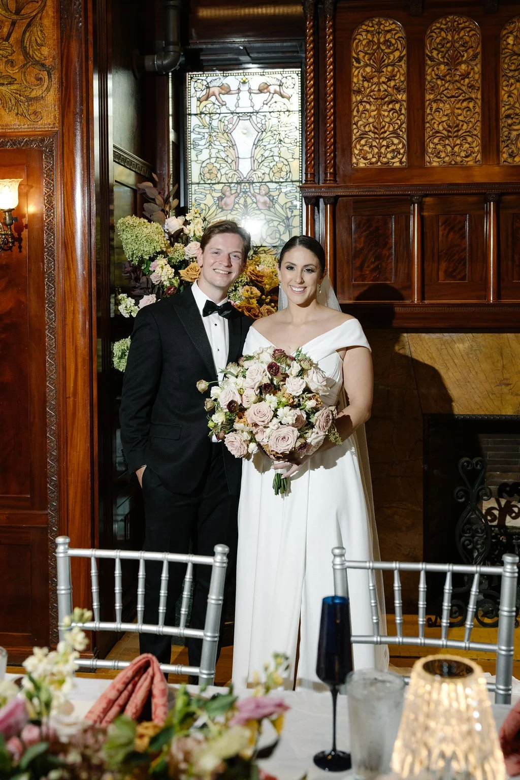 A bride and groom posing for a wedding photo indoors with wood-paneled walls, a stained glass window, and floral arrangements in the background. The bride is holding a bouquet of pink and white flowers, and the groom is wearing a black tuxedo with a 