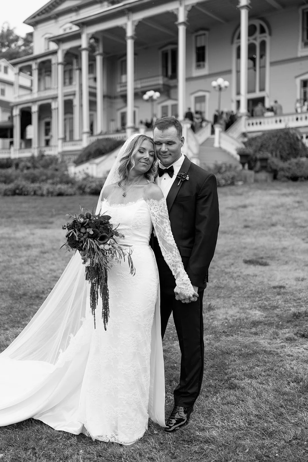 Black and white photograph of a bride and groom standing close together on a lawn in front of a large, ornate house. The bride is wearing a lace wedding gown and holding a bouquet, the groom is in a tuxedo with a bow tie. They are holding hands and g