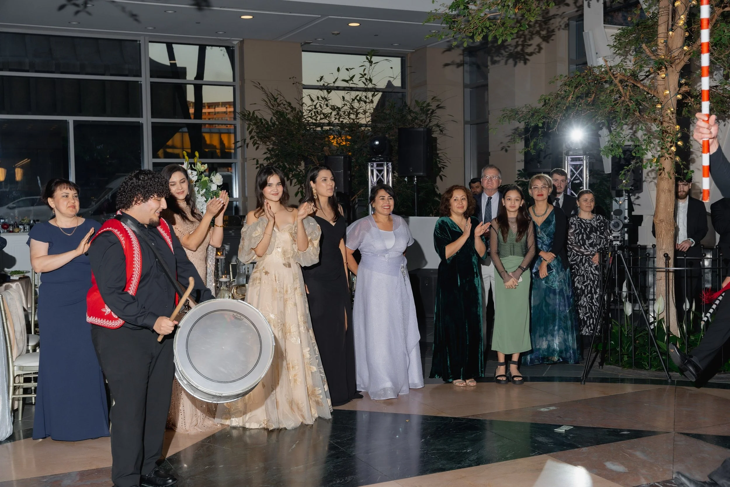 Group of people at a celebration, dressed in formal attire, with some clapping and others smiling, indoors with large windows and trees visible outside.