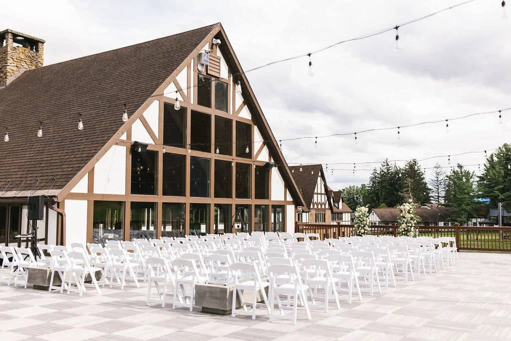 Outdoor wedding setup with white chairs and floral arrangements in front of a modern barn-style building with large windows and string lights overhead.