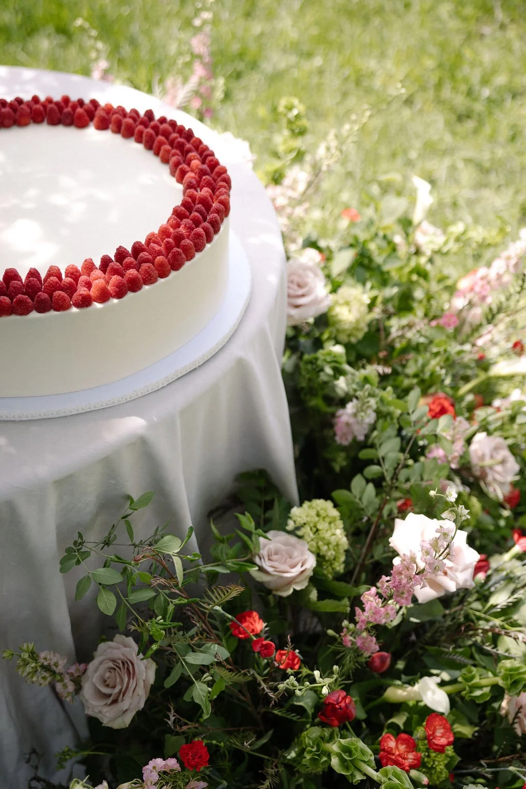 A white cake decorated with raspberries on a table covered with a white cloth, surrounded by various flowers including roses and other colorful blooms, outdoors with greenery in the background.