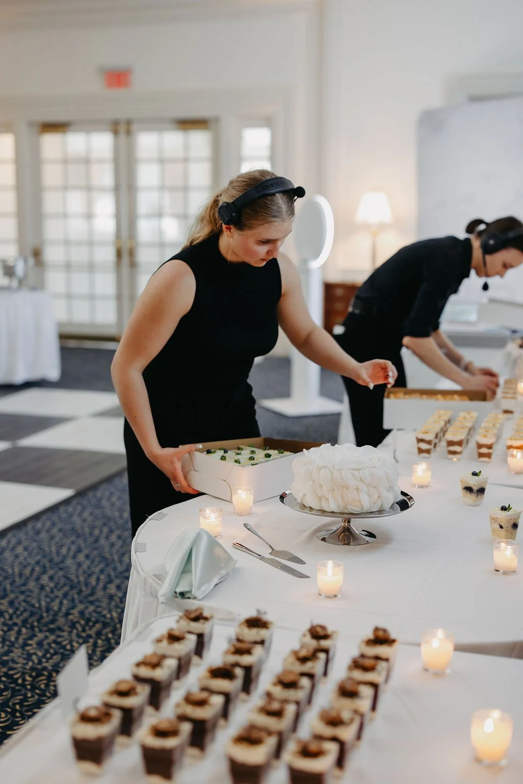 Two women in black dresses and black headsets preparing and arranging desserts on a table at a buffet-style event, with a variety of cakes, cupcakes, and snacks, surrounded by small lit candles.
