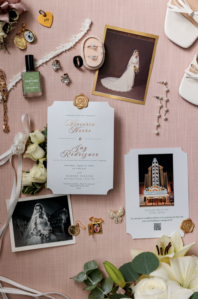 Wedding invitation surrounded by jewelry, flowers, photos, a perfume bottle, and various accessories on a pink surface.