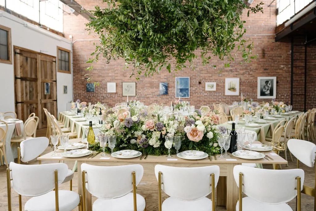 Set up for a formal event with a long table adorned with floral centerpieces, wine bottles, glasses, and plates in a decorated indoor venue with chairs, artwork on brick walls, and a hanging greenery ceiling.