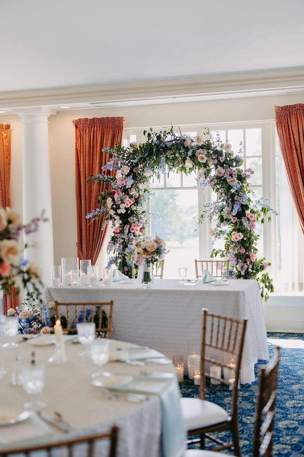 Wedding reception table with a floral arch, candles, and elegant table settings in a bright room with large windows and red curtains.